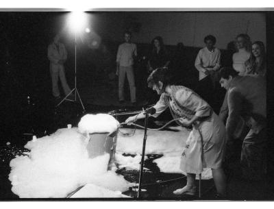 Audience members are gathered around Jane Ellison, feeding a tube into a plastic bin that is overflowing with bubbles.