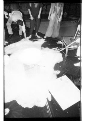 An overexposed shot of three people gathered around a plastic bin that is overflowing with bubbles. The liquid pours onto a tarp arranged on the floor. 