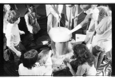 A group of people gather around a plastic garbage bin overflowing with bubbles. 