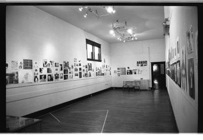 A wide-angle image from the east corner of a gallery space. There are a series of small pin-up images scattered across the walls. Two large light fixtures are mounted on the ceiling and illuminate the works. 
