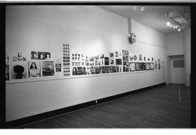 A wide-angle image captures a section of the gallery space where a collection of portraits in a variety of styles are mounted with pins on the wall. 
