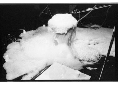 Soapy bubbles spill out of a plastic garbage can and overflow onto a tarp on the floor. A microphone is pointed towards the top of the bin. 