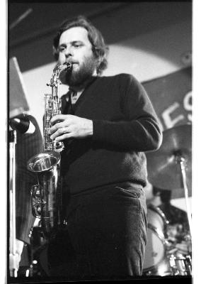 A musician plays the saxophone while wearing dark colors. Their hand and saxophone are captured in great focus, while everything else becomes blurred.