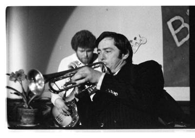  A musician playing the trumpet wears a black tuxedo and hair combed to the side. Behind them, is a musician playing a bass guitar next to potted daffodils.
