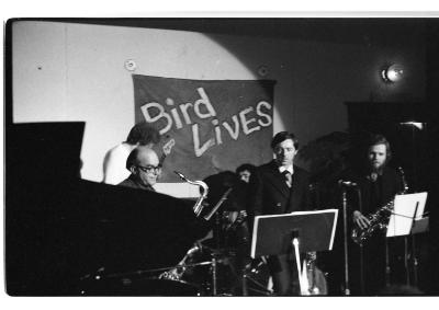 Five musicians are lit by a spotlight. They stand in two rows. The front row consists of a trumpeter in between two saxophonists, and in the back is a drummer and bassist. 