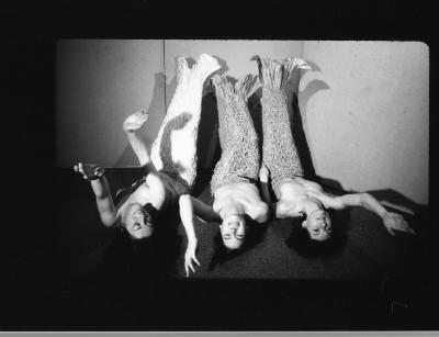 A black-and-white photograph of a television monitor showing three topless women wearing mermaid tails lying on the floor with their legs propped up against a wall.