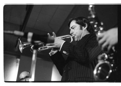 A blurry mic and saxophone occupy the foreground of the image. Just behind, in full focus, a trumpet player blows wind through their instrument while holding down the second button. 