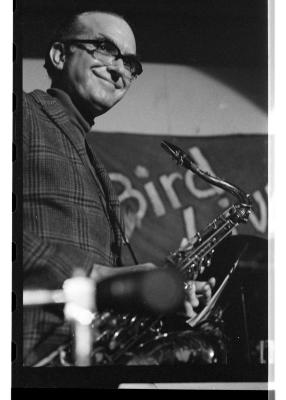 A saxophone player in a plaid suit smiles out at the crowd. Behind them you can see the curve of a spotlight and the painted phrase Bird.