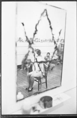 A mirror is scratched with an incomplete Star of David. In the reflection, Jane Ellison can be seen sitting nude on a chair, with audience members seated on the wooden floor around her.