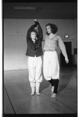 A black-and-white photo showing Susan McKenzie and Jennifer Mascall dancing. Susan’s arms are clasped above her head, and Jennifer holds her by the wrist. They both wear striped collared shirts, and leg warmers.