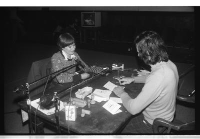 Hervé Fischer and Kate Craig sit across from each other at a wooden table positioned at the centre of the Grand Luxe Hall. Craig watches as Fischer stamps a piece of paper. The surface of the desk is cluttered with numerous rubber stamps, ink pads, papers, and pens. Two microphones on stands are angled towards their faces. 