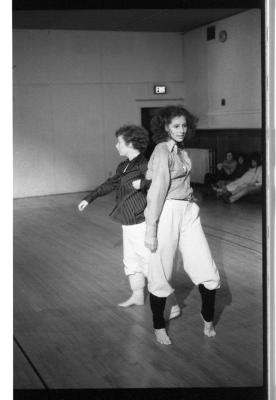 A black-and-white photo showing Susan McKenzie and Jennifer Mascall during a dance. They link arms, and both wear white pants, striped collared shirts, and leg warmers.