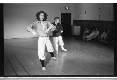 A black-and-white photo showing Jennifer Mescall and Susan McKenzie dancing. They wear collared shirts, white pants, and leg warmers.