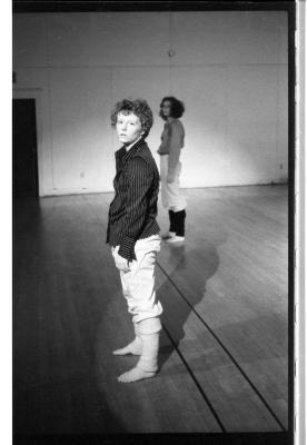 A black-and-white photo showing Susan McKenzie in profile, standing in a somewhat slouched pose. She wears a striped collared shirt, white pants, and leg warmers. Jennifer Mascall is behind her wearing a nearly identical outfit. 