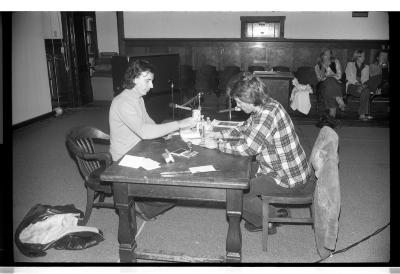 Hervé Fischer and Hank Bull sit across from each other at a wooden table positioned at the centre of the Grand Luxe Hall. Fischer stamps a piece of paper while Bull is writing on a separate piece of paper. The surface of the desk is cluttered with numerous rubber stamps, ink pads, pieces of paper , and pens. A jacket is strewn on the floor at the foot of Fischer’s chair, while Bull’s coat hangs on the back of his. Other audience members can be seen sitting along the periphery of the room.
