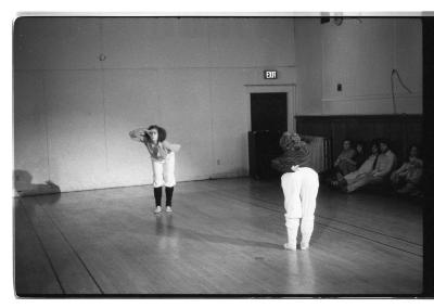 A black-and-white photo of Jennifer Mascall and Susan McKenzie facing each other while performing exaggerated seeking gestures. Leaning forward, they hold one hand above their brow, and the other behind their backs.