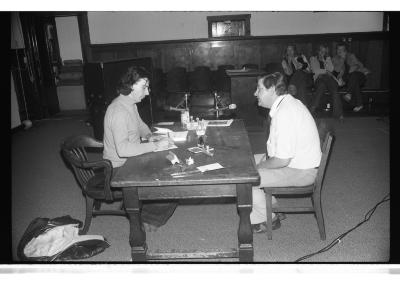 Hervé Fischer and Michael Morris sit across from each other at a wooden table positioned at the centre of the Grand Luxe Hall. Fischer is seen writing in a notebook while Morris speaks. A jacket is strewn on the floor at the foot of Fischer’s chair. The surface of the desk is cluttered with numerous rubber stamps, ink pads, papers, and pens. Other audience members can be seen seated along the periphery of the Grand Luxe Hall.