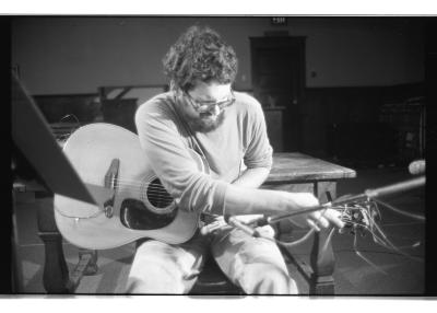 Eugene Chadbourne sits on a chair in the Grand Luxe Hall playing an acoustic guitar. Performing extended techniques, his hands grip the guitar’s neck.