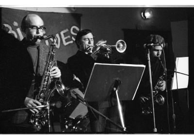 Two saxophonists lean forward, looking at sheet music on a stand. Meanwhile a trumpeter closes their eyes and leans back, playing with much force.