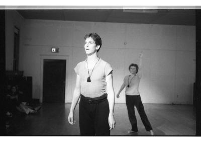 A black-and-white photo showing Seth Levinson standing with his arms by his sides. Behind him, Susan McKenzie lifts her left arm up towards the ceiling. They both wear t-shirts, dark pants, and matching necklaces with large pendants.