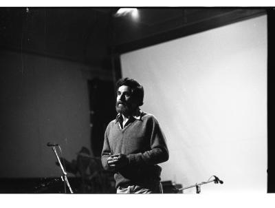 Allan Kaprow stands in the Grand Luxe Hall with his fingers clasped in front of his torso. He wears a v-neck sweater over top of a collared shirt.