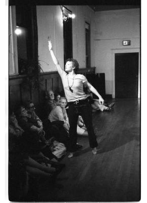 A black-and-white photo of Susan McKenzie dynamically reaching her left hand up to the ceiling. Audience members can be seen seated on the floor along the wall of the EDAM studio.