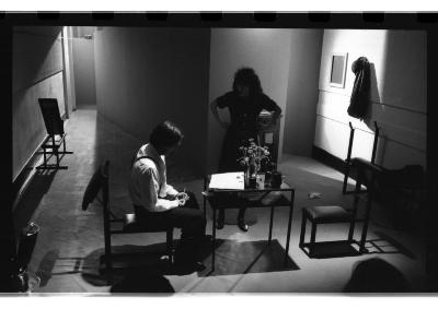 A triangular hallway opens up to a small table with seating for two. A performer dressed in a white shirt and suspenders slouches at the table as another performer dressed in a black talks down to them.
