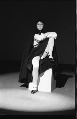 Lit by a spotlight, Jeanette Reinhardt sits on a plinth with one knee hugged into her chest. She wears a black cape that drapes over her head, a white blouse, a black skirt, stockings, and black patent Mary Janes. 