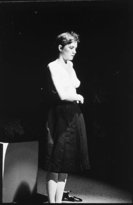 Jeanette Reinhadt stands under a spotlight. She is topless, and unbuttons the clasp on the side of her black pleated skirt.