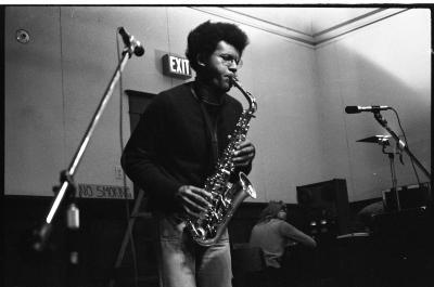 Leaning forward and holding down keys, the performer plays the saxophone. A hand-painted sign behind them reads "NO SMOKING".