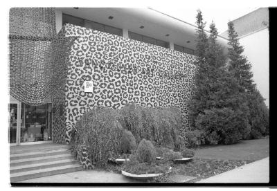 A black and white image documenting a leopard print mural by Eric Metcalfe and Kate Craig that spans the facade of the Vancouver Art Gallery. A canopy made from woven video tapes by Evelyn Roth hands above the stairs at the entrance to the gallery. 