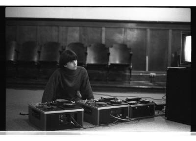 Lisa Steele sits on the floor of the Grand Luxe Hall behind three reel to reel tape machines. A video monitor is propped on a plinth and the  screen casts a white glow. 