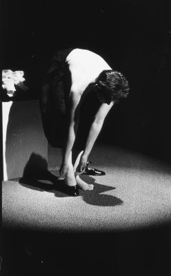 Jeanette Reinhardt is lit by a spotlight as she bends over to unbuckle her Mary Janes. She is topless and wears a black pleated skirt. 