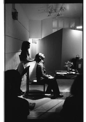 A performer sits at a small dining table tilting their head back to be cushioned by the hands of another actor wearing an apron. A light shines on the scene, illuminating the performers and darkening two onlookers into silhouettes. 
