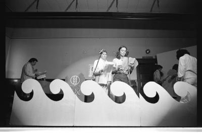 Two performers stand behind microphones holding pamphlets. A spotlight beams down on them. Four other artists read their scripts in the shadows. 