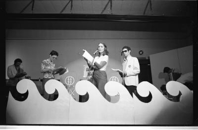 Performers arrange themselves to face an artist at center stage holding up a booklet. The artist speaks into a mic and glances to the back of the room.
