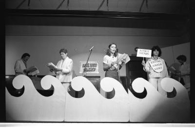 Artists holding booklets of paper stand behind flat prop waves that reach just below their hips. On the wall, is a taped sign reading LUX RADIO PLAYERS. One performer speaks into a microphone. Another wears a paper bib around their neck which reads ENTIRE WHALE ARMY, while holding up a sign that says ATTACK SOUNDS.