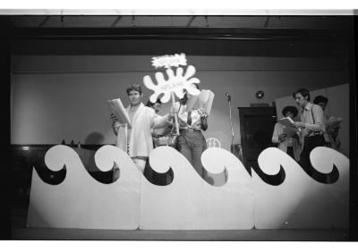 A performer holds up an expressive sign reading SPLASH. They step in front of an artist reading into a mic and reach a fence of prop waves.