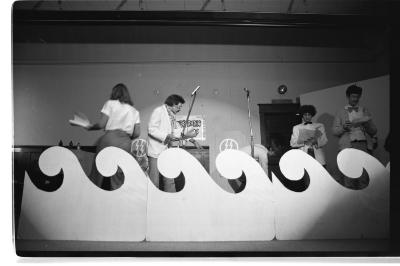 Four people, some in suit jackets and bow ties, read from scripts while standing behind a large wave prop on a stage. The image is in black and white.