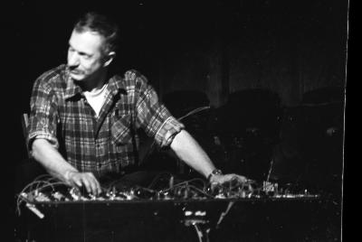 A ¾ medium-long shot of a man in a plaid shirt, as he extends both his arms out to adjust the knobs on a mixer console. The image is in black and white. 