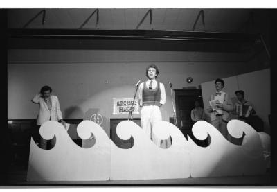 A play unfolds on a stage demarcated by a large prop in the shape of a curling wave. Someone wearing a collared shirt, tie, and knit vest is spotlit at centre-stage. He speaks into a microphone while holding his hands behind his back. Other actors and an accordion player can be seen in the wings. A sign that reads Lux Radio Players is taped to the wall behind the actors. The image is in black and white.
