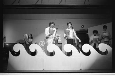 Two performers stand behind a minimal set of waves while holding rounded props. They focus on the papers in their hands, awaiting action.