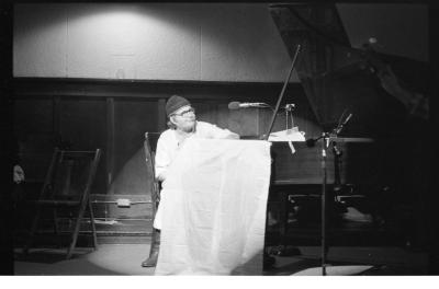 A man wearing a light coloured tunic and pants, round glasses, and a toque plays grand piano. A piece of cloth is wrapped over the side of the piano. This image is in black and white.