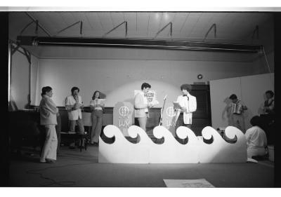 Performers gather near the back wall of a room between scattered furniture and props. Two performers stand in conversation under a spotlight, while another prepares to play an accordion.