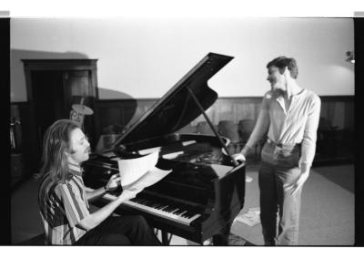 Hank Bull sits at a piano, smiling while flipping through sheet music. Pascal stands next to the piano with a coquette smile. The image is in black-and-white.