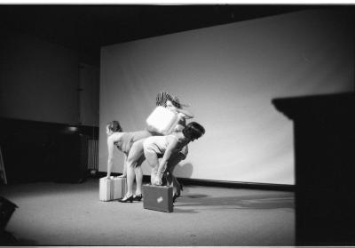 Two women wearing heels and bathing suits bend over at the waist, back-to-back. They create a makeshift table for a third woman, wearing a zebra-print sunhat, to place a white suitcase on. The two women each use a suitcase for balance.
