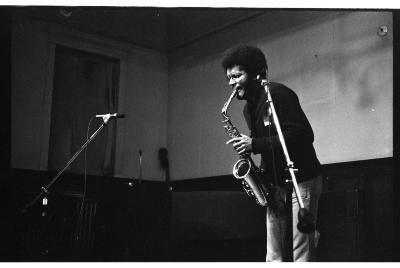 The musician takes a breath with the saxophone’s mouthpiece resting between their teeth. They lean forward with their left hand lightly holding the instrument's valves. 