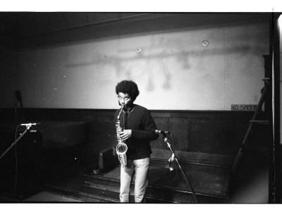 Standing below wooden stage steps, the artist plays their saxophone and glances up to the ceiling. Shadows on the wall behind them reveal a beam holding many cables.