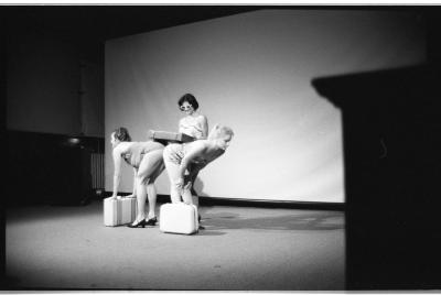 Two women wear heels and bathing suits, bending over at the waist with their backs to each other. Their backs create a table for a third woman, who wears a bathing suit and sunglasses, to place a suitcase on. The woman on the left places her hands on a suitcase for balance, while the one on the right has her hands on her knees and a suitcase by her side. 
