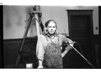The author stands in front of a triangular wooden structure with one hand on a podium and the other on their hip. They look straight into the camera with little expression.